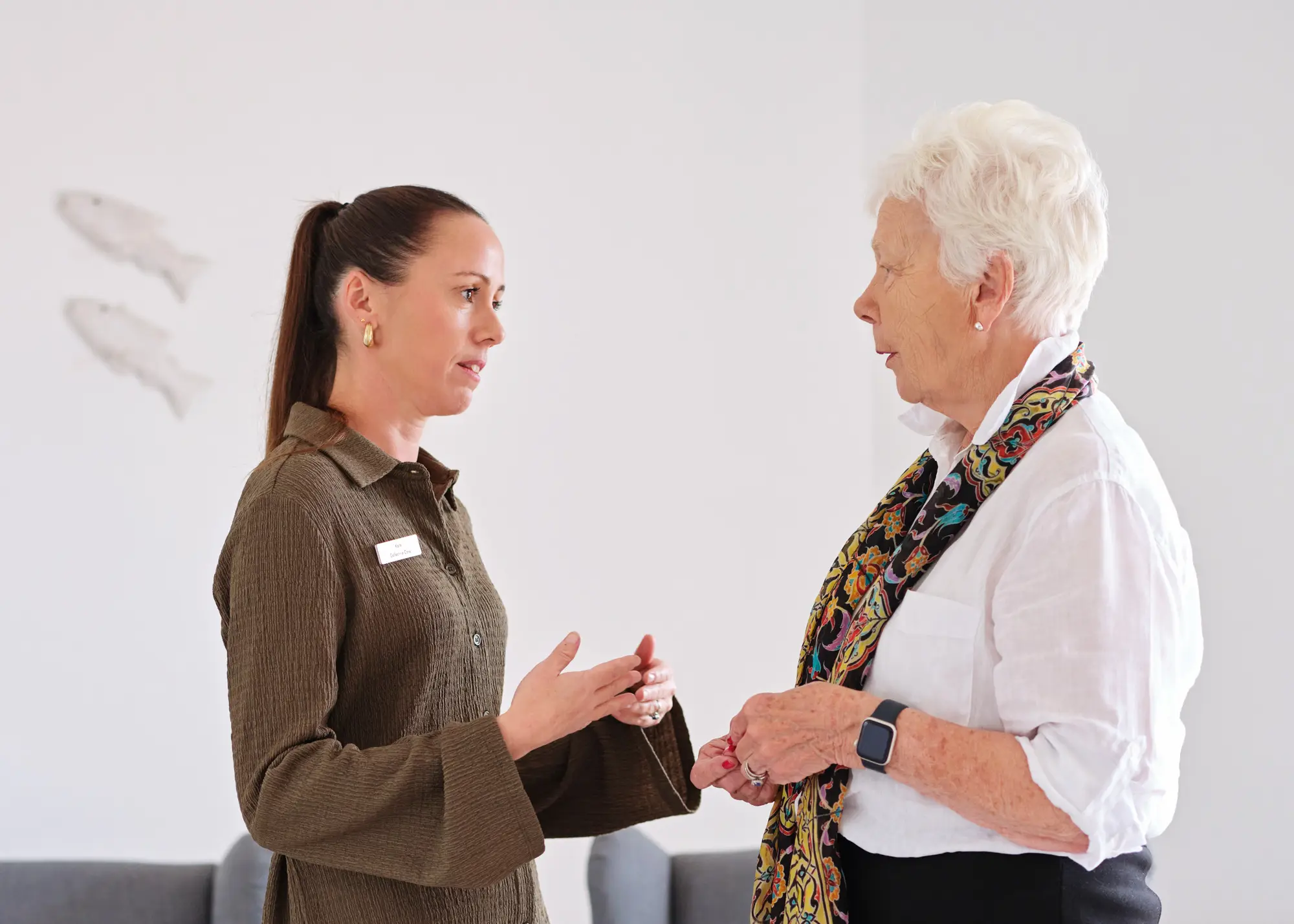A carer talking attentively with an older woman as she stands supported at home.