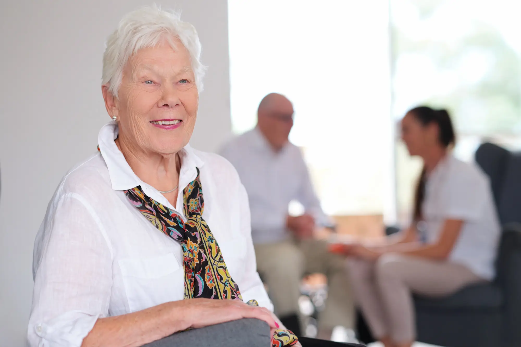 An older woman smiling warmly at home, with a carer and family member chatting in the background.